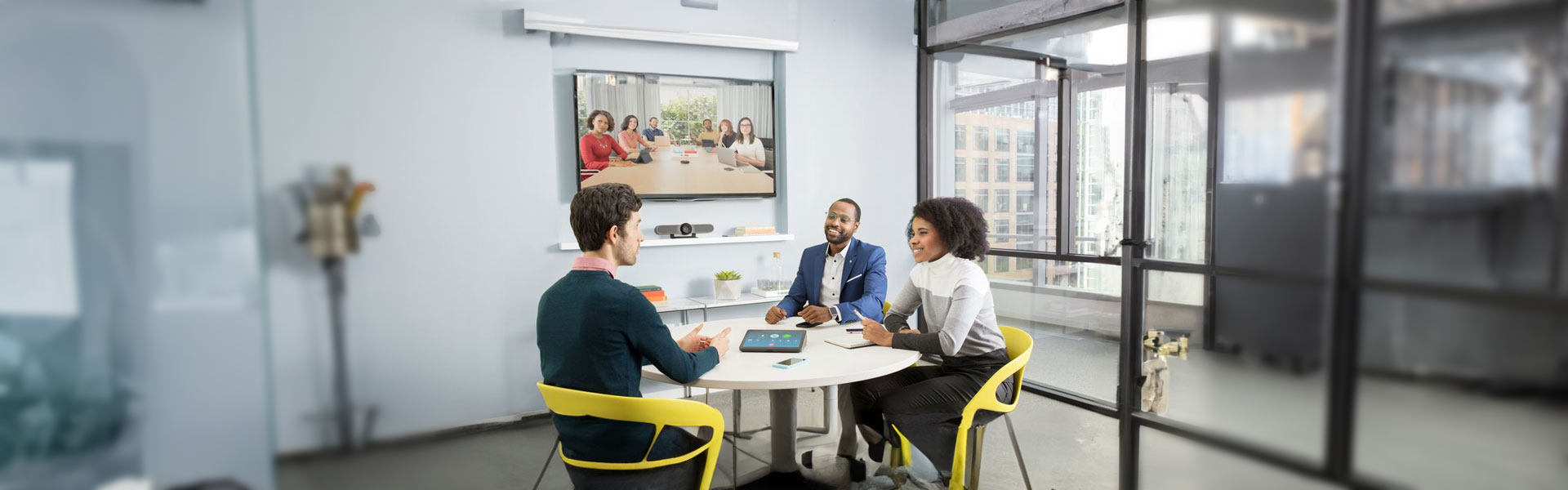 Tres personas en una sala pequeña frente a una pantalla con videollamada y panel de control sobre la mesa